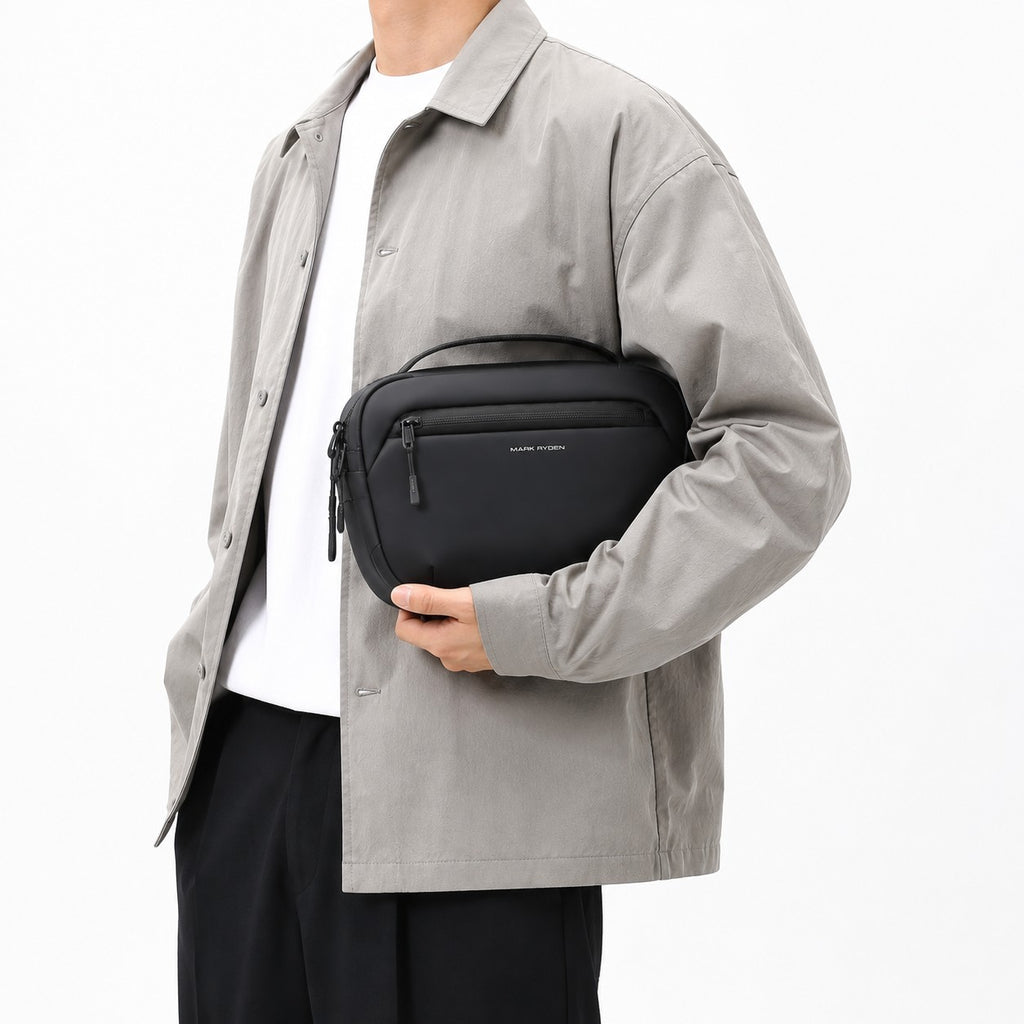 Sac bandoulière noir Mark Ryden pour homme, compact et imperméable, porté à la main sur un fond blanc.
