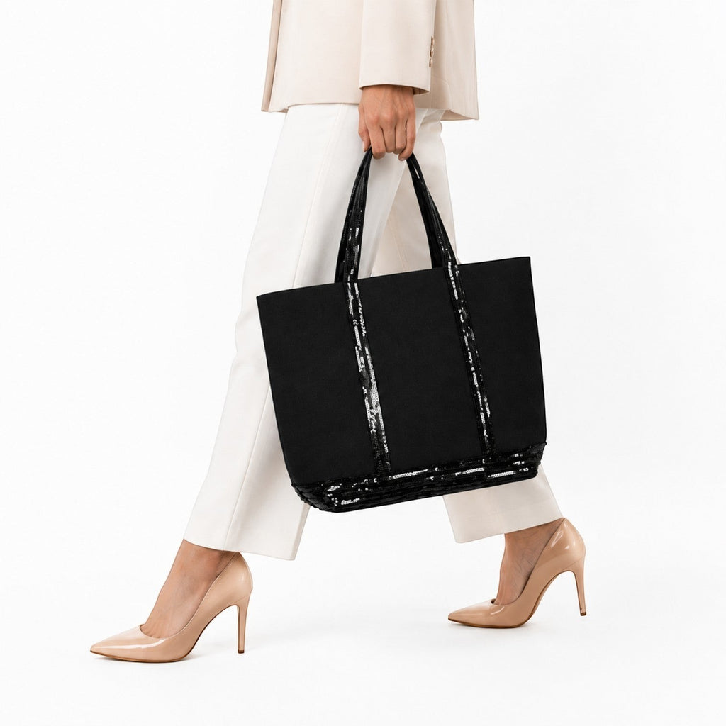 Sac fourre-tout noir en toile avec garniture à paillettes, anses et bordure, porté par une femme en tenue de bureau.