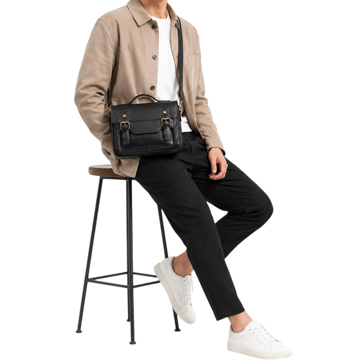 Homme portant un sac messager Univers Sac en cuir noir style vintage avec bandoulière, assis sur un tabouret